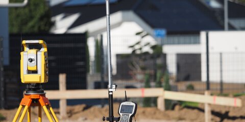 Gelbes Vermessungsgerät auf einem Stativ neben einer Messlatte mit angebrachtem Steuergerät. Im Hintergrund unscharf ein modernes Gebäude und eine Baustelle.