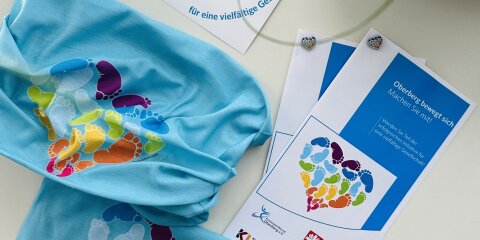 Ein Flyer, ein Tuch und eine Karte mit bunten Fußabdrücken in Herzform liegen auf einem Tisch. Rechts steht eine Holzschale, links ist eine Pflanze sichtbar.