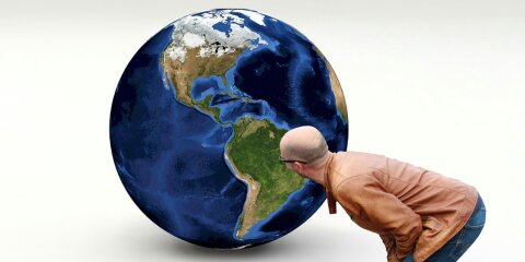 Ein Mann in einer braunen Lederjacke und Jeans beugt sich vor, um eine große Erdkugel mit Ansicht der westlichen Hemisphäre genauer zu betrachten. Hintergrund ist hell und neutral.