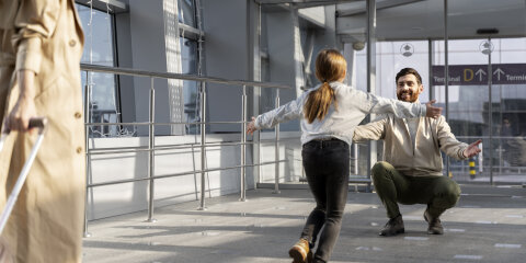 Ein Mann in heller Kleidung hockt lächelnd mit ausgebreiteten Armen in einer Flughafenhalle, während ein Mädchen mit langen Haaren auf ihn zuläuft. Im Vordergrund ist eine Person mit beigem Mantel und Koffer zu sehen.