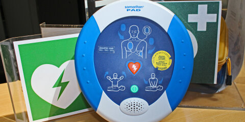 Ein automatisierter externer Defibrillator (AED) der Marke Samaritan PAD auf einem Tisch. Im Hintergrund sind grüne Symbole mit Herz und Kreuz sichtbar.