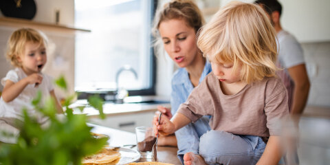 Eine Frau und zwei Kinder in einer Küche. Ein Kind sitzt auf der Arbeitsfläche und bestreicht einen Pfannkuchen, während das andere einen Löffel hält. Im Hintergrund ein Mann und eine Pflanze im Vordergrund.