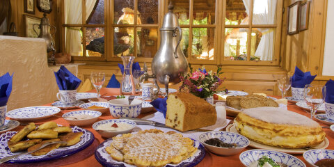 Ein gedeckter Tisch mit traditionellen Speisen wie Waffeln, Brot und gefüllter Pfannkuchen, ein blau-weißes Kaffeeservice, einer silbernen Kanne (Dröppelminna) und einem Blumenstrauß in einem Restaurant vor einem Holzfenster.
