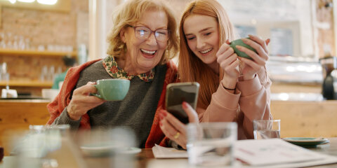 Happy grandmother and her adolescent granddaughter sitting in a cafe, enjoying coffee and using smart phone for funny videos.