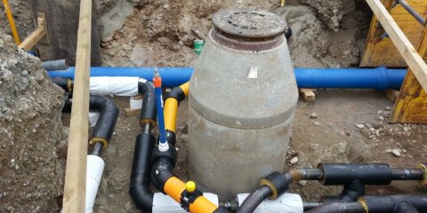 Baugrube mit freigelegten Rohren unterschiedlicher Größe und Farben sowie einem Betonschachtdeckel. Holz- und Metallelemente zur Abstützung der Grube.