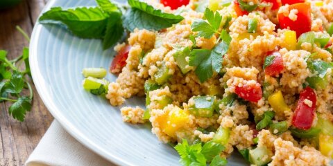 Ein Teller mit Couscous-Salat, garniert mit frischen Kräutern. Der Salat enthält bunte Paprikastücke, Gurken und Tomaten. Der Teller steht auf einem Holzuntergrund.