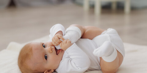 Ein Baby liegt auf dem Rücken auf einer weichen Decke, trägt einen weißen Strampler und weiße Söckchen und hält einen Fuß mit den Händen.