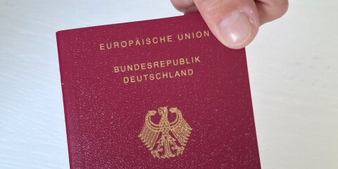 Zwei Hände halten gemeinsam einen deutschen Reisepass mit bordeauxrotem Einband und goldener Prägung, auf der der Bundesadler zu sehen ist.