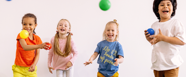 Vier Kinder spielen mit bunten Bällen vor einem weißen Hintergrund. Sie tragen bunte Kleidung und sind in Bewegung.