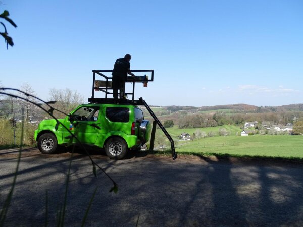 Eine Person steht auf einer Plattform, die auf einem grünen Geländewagen montiert ist. Der Wagen steht auf einer Straße mit Blick auf eine ländliche Landschaft mit Feldern und kleinen Häusern. Die Person reinigt einen Wegweiser für den Radverkehr.