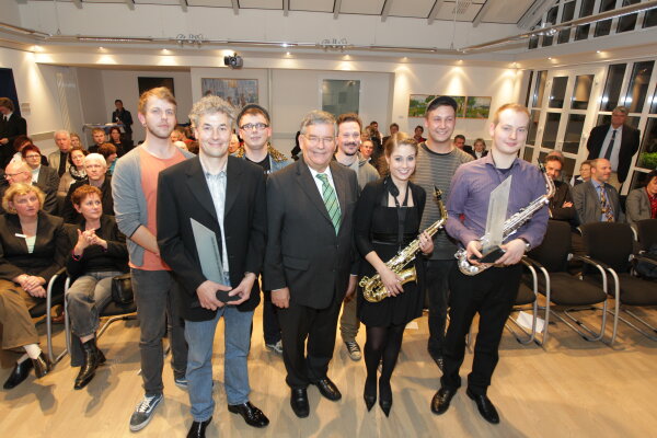 Eine Gruppe von Personen steht in einem Veranstaltungsraum. Zwei Personen halten Saxophone, eine weitere eine Auszeichnung. Im Hintergrund sitzen Zuschauer.
