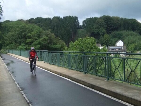 Eine Person mit Helm und roter Jacke fährt ein Fahrrad auf einem nassen Weg mit einem grünen Geländer und Bäumen im Hintergrund.