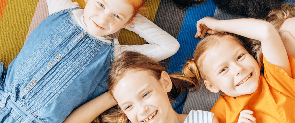 Vier Kinder liegen auf einem bunten Streifenteppich. Ihre Gesichter sind unkenntlich gemacht. Ein Kind trägt ein Jeanskleid, ein anderes ein orangefarbenes Oberteil.