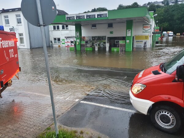 Eine überflutete Tankstelle mit Wasser, das bis zu den Zapfsäulen reicht. Zwei rote Feuerwehrfahrzeuge sind am Straßenrand sichtbar.
