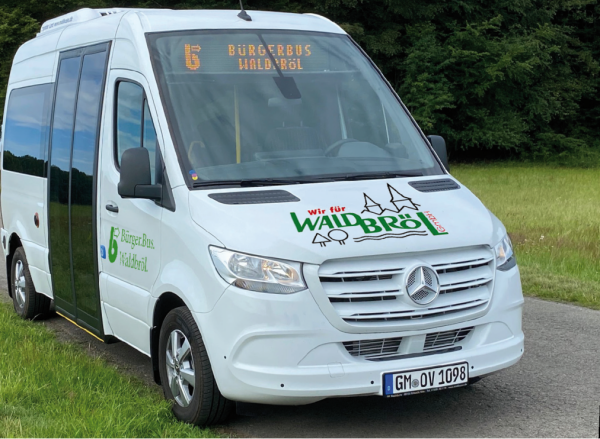 Ein weißer Kleinbus mit der Aufschrift 'Bürgerbus Waldbröl' und einem Mercedes-Logo steht auf einer Straße vor einer Wiese und Bäumen.