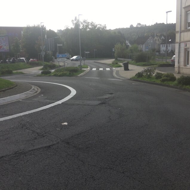 Eine Straßenszene mit einem Kreisverkehr, Zebrastreifen und umliegenden Grünflächen. Rechts steht ein Gebäude mit einem Schild für Thai-Massage und Wellness.