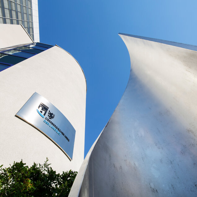 Moderne Gebäudefassade mit gebogenem Design, Metallskulptur im Vordergrund und blauem Himmel im Hintergrund. Ein Schild zeigt ein Wappen und Text.