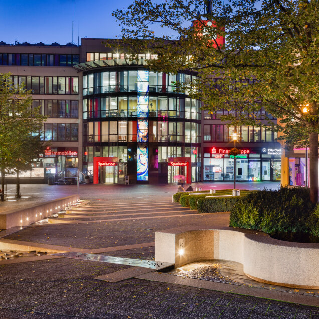 Städtischer Platz bei Nacht mit moderner Architektur, beleuchteten Gebäuden, einem kleinen Wasserlauf, Sitzgelegenheiten und Bäumen im Vordergrund.