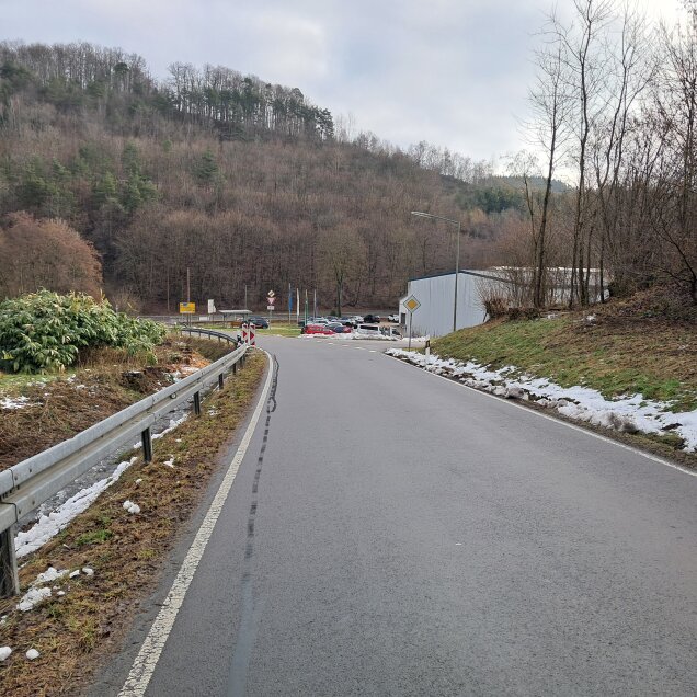 Eine kurvige Straße mit Leitplanken führt durch eine ländliche, bewaldete Gegend. Auf beiden Seiten der Straße liegt teils geschmolzener Schnee. Im Hintergrund sind ein Parkplatz, ein Gebäude und weitere Bäume erkennbar.
