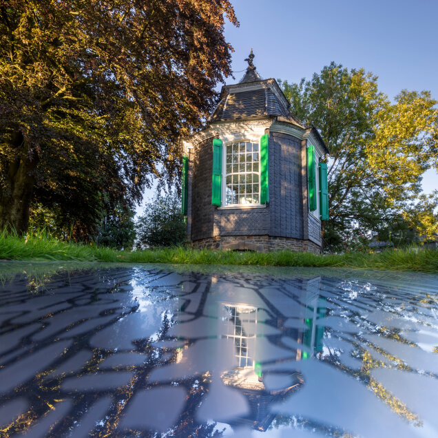 Kleines achteckiges Gebäude mit grünen Fensterläden, umgeben von Bäumen und Gras. Die Struktur spiegelt sich in einer glatten Oberfläche im Vordergrund.