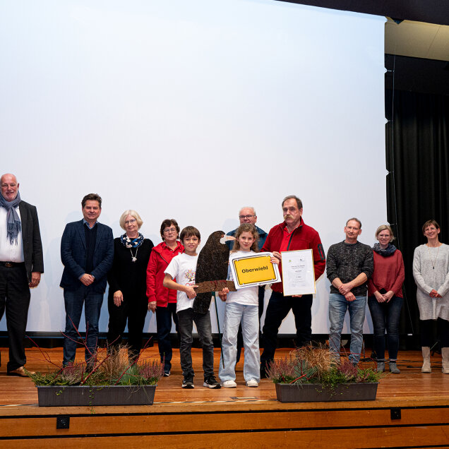 Eine Gruppe von Personen steht auf einer Bühne vor einer weißen Leinwand. Einige halten ein Schild und eine Urkunde. Vorne auf der Bühne stehen Blumenkästen.
