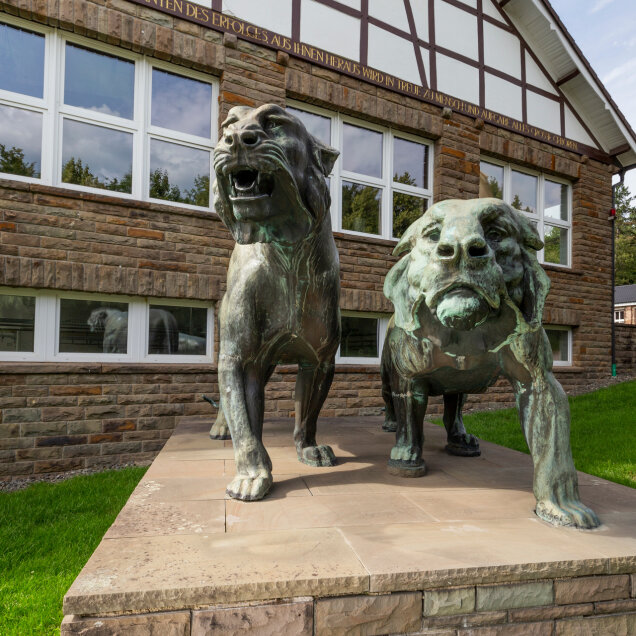 Zwei bronzene Löwenstatuen auf einem Sockel vor einem Gebäude mit Fachwerk und Steinfassade. Daneben eine Fahnenstange und gepflegter Rasen.