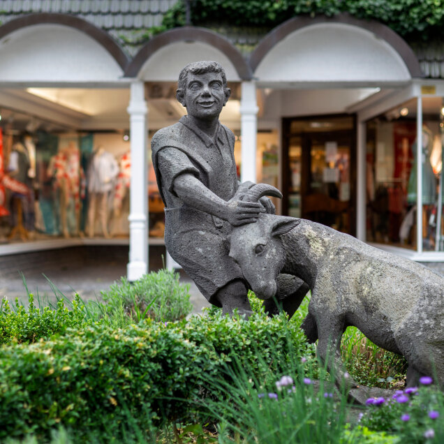 Steinfigur eines Jungen, der eine Ziege am Kopf hält, umgeben von Pflanzen. Im Hintergrund ein Geschäft mit Schaufenstern und Bekleidungsauslage.