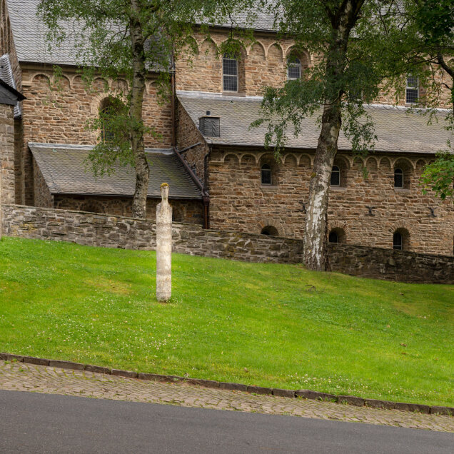 Historisches Gebäude aus Sandstein mit Rundbogenfenstern, umgeben von einer Steinmauer. Vor dem Gebäude stehen drei Steinsäulen auf einer grünen Wiese, flankiert von Bäumen.