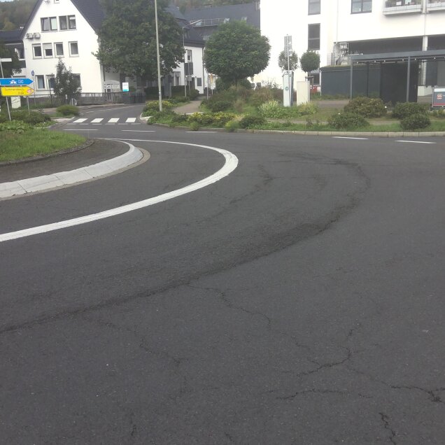Eine Straßenkurve mit Fahrbahnmarkierungen, umgeben von Grünflächen und Büschen. Im Hintergrund stehen Wohnhäuser und ein Parkplatz.