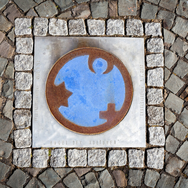 Eine quadratische Metallplatte mit einem Symbol in der Mitte, das eine blaue und rostfarbene Darstellung zeigt. Umgeben von Kopfsteinpflaster.