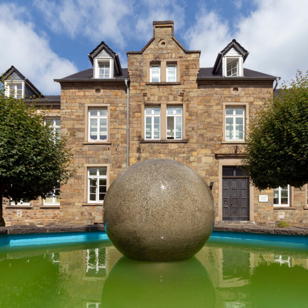 Ein steinernes Gebäude mit heller Fassade und weißen Fenstern. Im Vordergrund ein Brunnen mit grünem Wasser und einer großen, runden Steinskulptur. Umgeben von Bäumen und blauem Himmel.