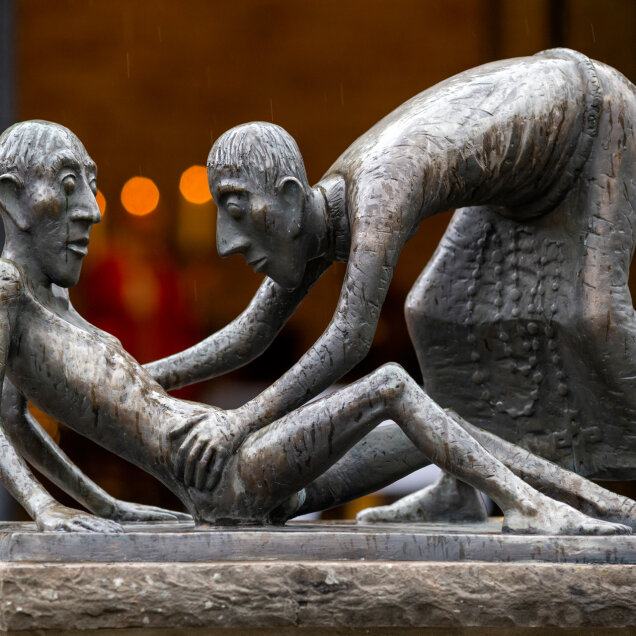 Bronzestatue zweier Figuren: eine stehende Person beugt sich über eine sitzende, hager wirkende Person. Beide haben stilisierte Gesichtszüge und große Augen.