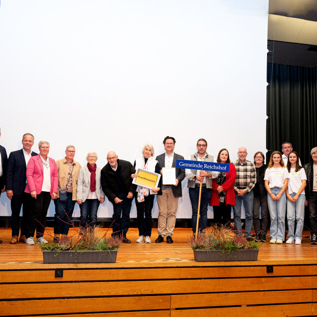 Eine Gruppe von Menschen steht auf einer Bühne vor einer weißen Leinwand. Einige halten Schilder und Dokumente. Im Vordergrund sind Blumenkästen.