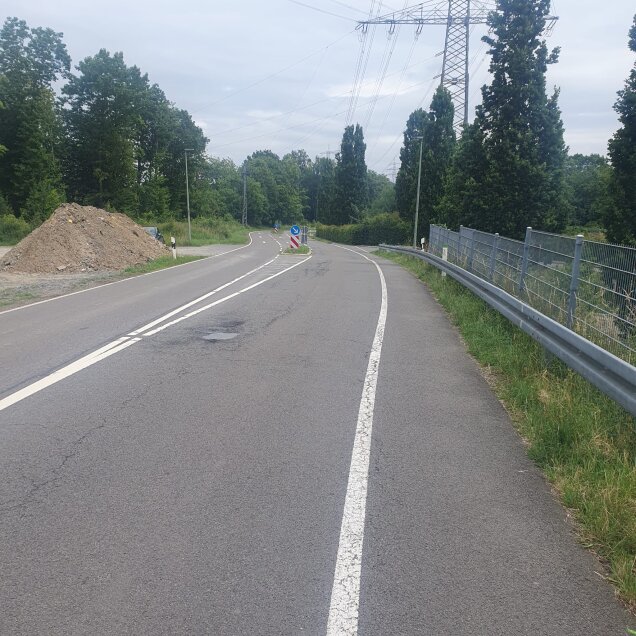 Eine kurvige Landstraße mit durchgezogener und gestrichelter Mittellinie, rechts ein Metallzaun und links ein Erdhaufen. Im Hintergrund Bäume und Strommasten.