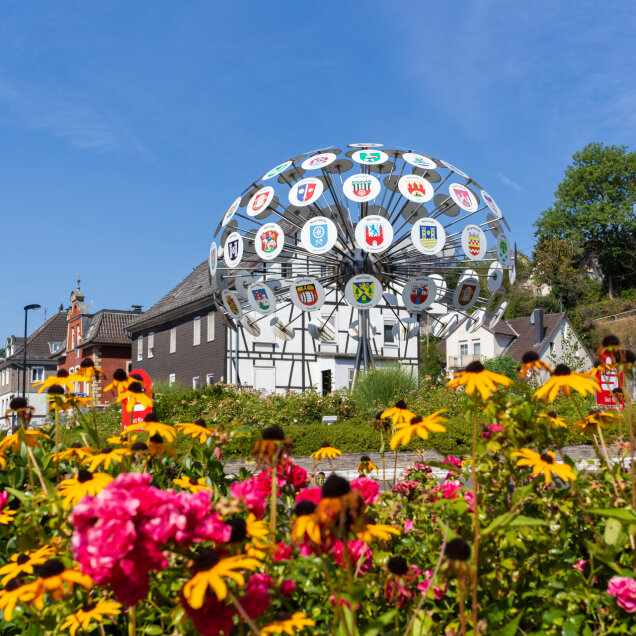 Eine große Metallskulptur mit Wappen verschiedener Städte steht vor Wohnhäusern. Im Vordergrund sind gelbe und pinke Blumen zu sehen. Der Himmel ist blau.