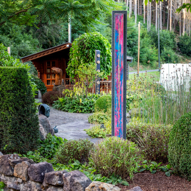 Ein gepflegter Garten mit einer Steinmauer, Sträuchern, einem modernen Kunstobjekt in bunten Farben, einer Statue und einem kleinen Holzhaus im Hintergrund.