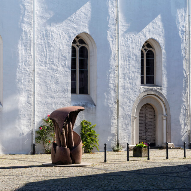 Weiße Kirchenfassade mit gotischen Fenstern und Türen, davor eine moderne Skulptur aus Metall, Blumenbeete und Pflastersteine. Sonnenlicht und Schatten sichtbar.