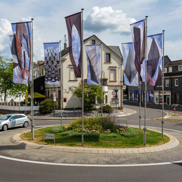 Kreisverkehr mit bepflanzter Mitte, umgeben von mehreren Fahnenmasten mit dekorativen Fahnen. Eine Radfahrerin und ein Auto sind auf der Straße sichtbar. Im Hintergrund Gebäude und Geschäfte.