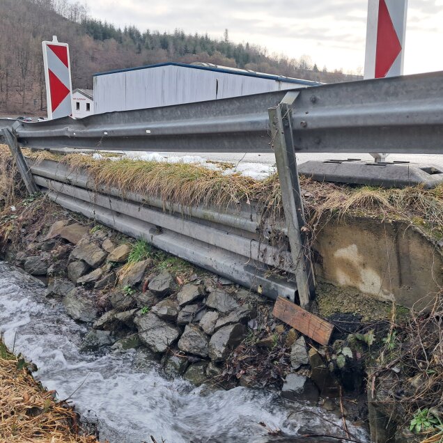 Ein beschädigtes Straßengeländer aus Metall über einem fließenden Bach, mit steinigem Ufer und Vegetation. Im Hintergrund ein Gebäude und Bäume.