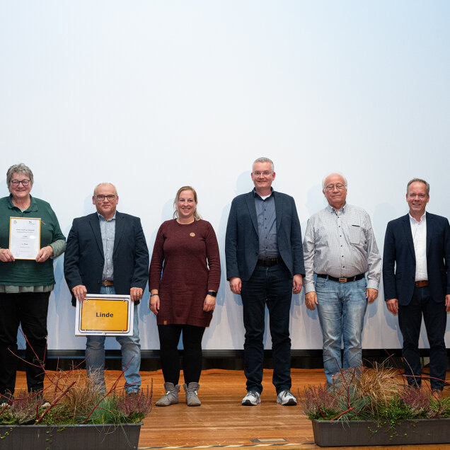 Eine Gruppe von acht Personen steht auf einer Bühne vor einer weißen Leinwand. Zwei Personen halten Schilder, eines mit dem Wort 'Linde', das andere scheint eine Urkunde zu sein. Im Vordergrund sind Blumenkästen mit Pflanzen sichtbar.