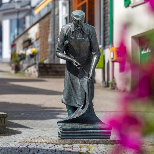 Bronzestatue eines Mannes in Arbeitskleidung, der ein Stück Stoff hält, auf einem Pflasterweg in einer Stadt mit umliegenden Gebäuden und Blumen im Vordergrund.