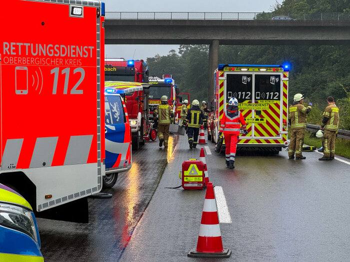Einsatzfahrzeuge und Rettungskräfte auf einer nassen Straße, darunter Rettungswagen, Feuerwehrfahrzeuge und Personen in Einsatzkleidung. Verkehrshütchen sichern den Bereich ab, im Hintergrund ist eine Brücke sichtbar.