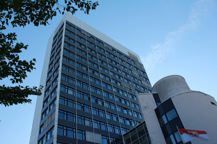 Ein modernes Hochhaus mit vielen Fenstern vor einem blauen Himmel. Im Vordergrund ein kleineres, rundes Gebäude und ein Baumzweig.