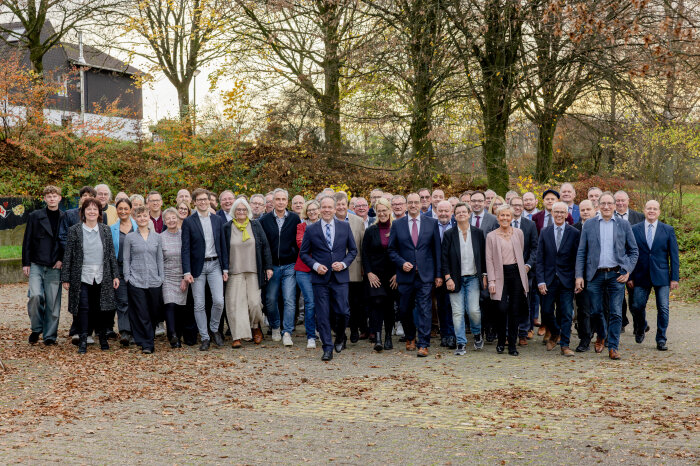Eine große Gruppe von Menschen in herbstlicher Umgebung, einige in Businesskleidung, andere in Freizeitkleidung. Im Hintergrund sind Bäume und ein Gebäude sichtbar.