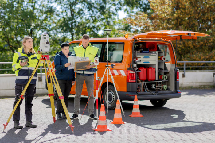 Drei Personen in Sicherheitskleidung arbeiten mit Vermessungsgeräten vor einem orangefarbenen Fahrzeug mit geöffnetem Kofferraum auf einem Parkplatz.