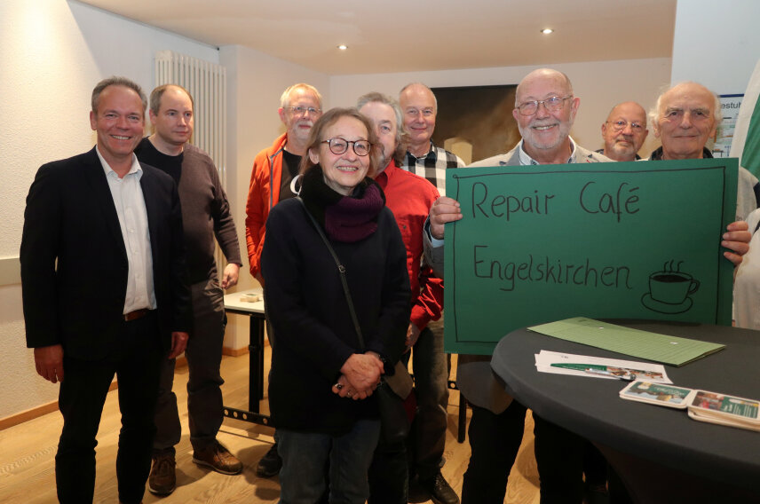 Eine Gruppe von Personen steht in einem Raum und lächelt. Eine Person hält ein grünes Schild mit der Aufschrift 'Repair Café Engelskirchen' und einer gezeichneten Kaffeetasse.