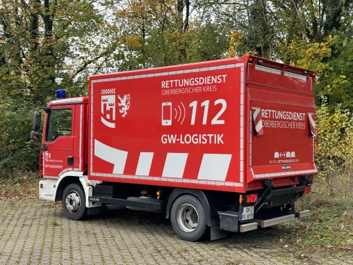 Rotes Einsatzfahrzeug mit der Aufschrift 'Rettungsdienst Oberbergischer Kreis' und '112 GW-Logistik', geparkt auf einem gepflasterten Platz, umgeben von Bäumen im Herbst.