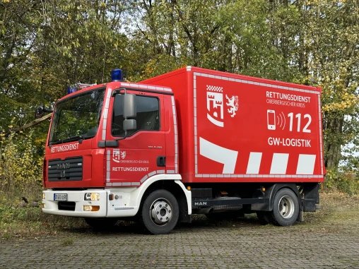 Ein rotes Einsatzfahrzeug des Rettungsdienstes Oberbergischer Kreis, beschriftet mit 'GW-Logistik' und Notrufnummer 112, steht auf gepflastertem Untergrund vor Bäumen.