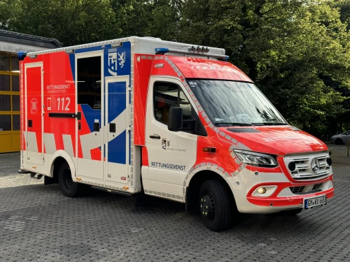 Ein Rettungswagen des Oberbergischen Kreises in Deutschland, rot-weiß-blau gestaltet, steht auf einem gepflasterten Parkplatz vor einem Gebäude mit gelben Toren.