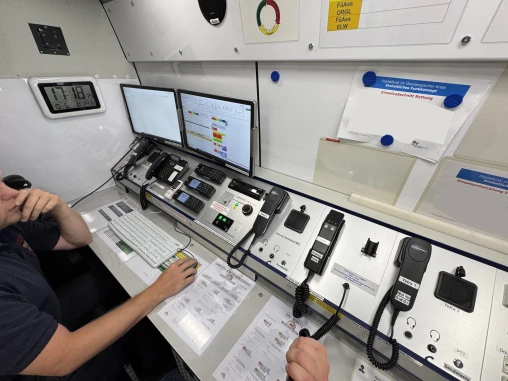 Arbeitsplatz in einer Einsatzleitstelle mit zwei Monitoren, Kommunikationsgeräten wie Funksprechgeräten und Telefonen, sowie einer Tastatur. Eine Person sitzt am Schreibtisch.
